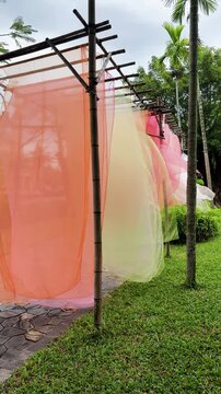 Vibrant outdoor art installation made of translucent pink, orange, yellow, green and white fabrics flowing in wind along landscaped park walkway, shot handheld, vertical video. High quality 4k footage