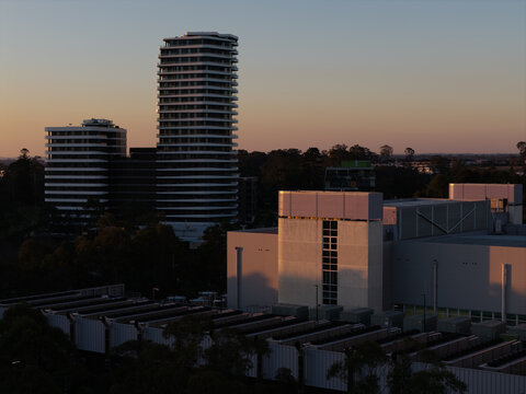 Aerial view of a modern data center facility with cooling units on the roof, adjacent to high-rise residential apartment buildings during golden hour in Sydney, New South Wales, Australia.
