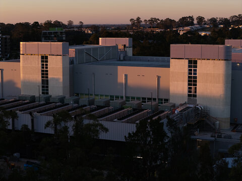 Aerial view of a modern data center facility at golden hour, featuring rooftop cooling infrastructure, surrounding trees, and suburban landscape in Sydney, New South Wales, Australia.
