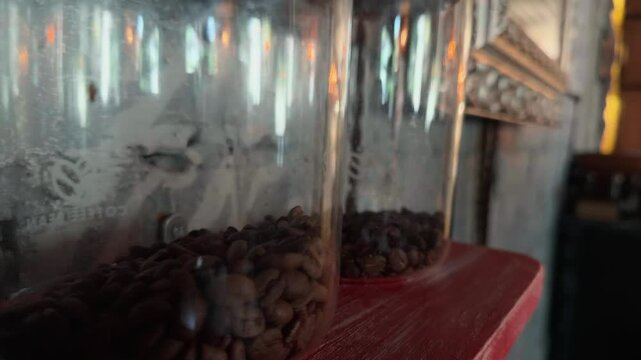 Atmospheric Close Up Dolly Shot Sliding Past Glass Jars of Dark Roasted Whole Coffee Beans Standing in a Row on Scratched Red Wooden Shelf in Industrial Loft Cafe Rough Concrete Wall Moody Ambience