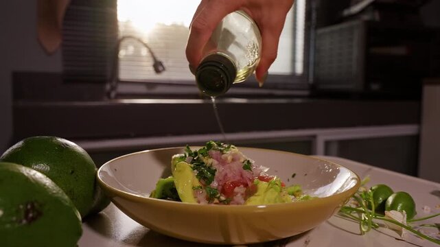 Woman Pours Cooking Oil into Bowl of Avocado, Tomato, Onion and Cilantro, Preparing Fresh Homemade Guacamole in Sunlit Kitchen