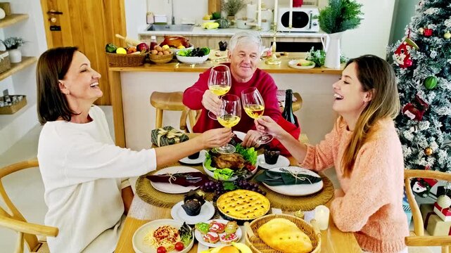 Three people toast with white wine around a festive holiday dinner in a cozy kitchen, with a decorated Christmas tree and plenty of homemade dishes