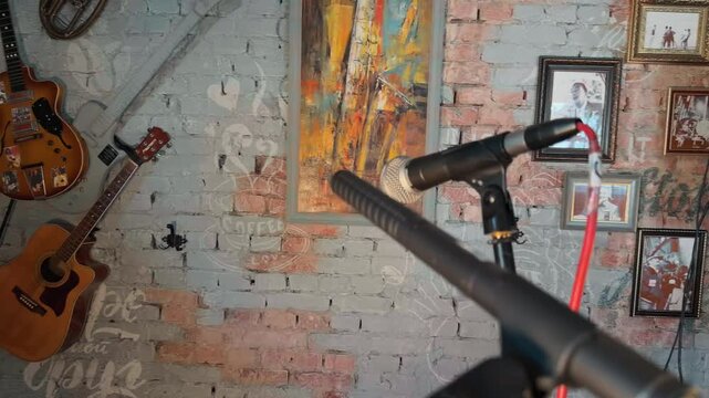 Professional broadcasting microphones on stands with red audio cable in foreground inside vintage recording studio with painted brick wall decorated by old art and acoustic guitars handheld arc shot