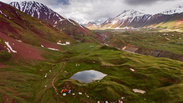 Aerial view of Tulpar Lake surrounded by lush green rolling hills, snow-capped mountains, and dramatic red slopes under a cloudy sky in Alay District, Osh Region, Kyrgyzstan.