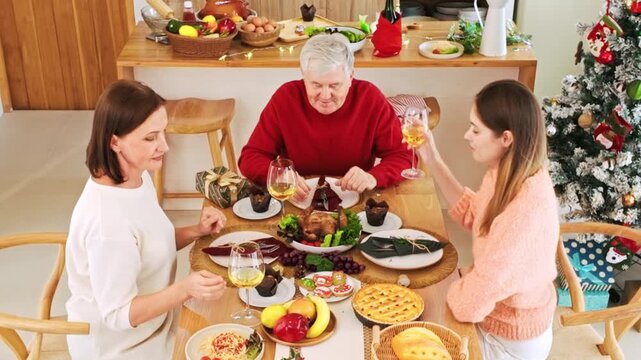 Three people toast with white wine around a festive holiday dinner in a cozy kitchen, with a decorated Christmas tree and plenty of homemade dishes