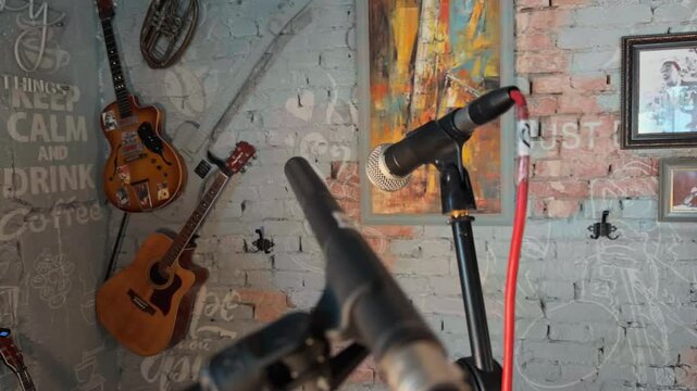 Professional microphones on stands ready for live music performance on empty stage inside cozy retro comedy club with painted brick wall vintage guitars and neon sign handheld orbiting arc shot