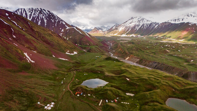 Aerial view of Tulpar Lake surrounded by lush green meadows, rolling hills, and snow-capped mountains under a cloudy sky in Alay District, Osh Region, Kyrgyzstan.