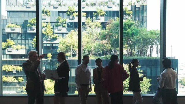 Silhouetted professionals networking by office windows, overlooking a modern green building with lush planted terraces in the city