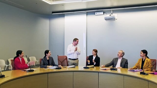 A team in a boardroom applauds a standing colleague during a business meeting, with microphones and a projector in a modern conference setting.