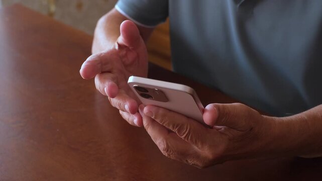 Close-up view of an older adult's hands using a modern smartphone, scrolling through the screen and sending a text message while sitting at a wooden table in a dimly lit room