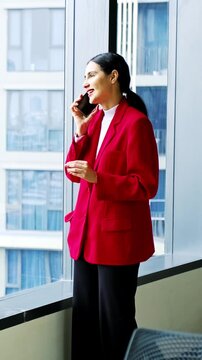 A professional in a red blazer talks on a smartphone by large office windows, smiling as city high-rises frame the bright, modern workspace