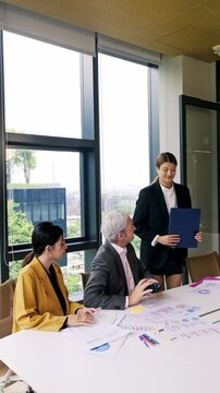 Senior businessman reviews financial reports and charts with two female colleagues during a focused team meeting in a bright modern office with city views.