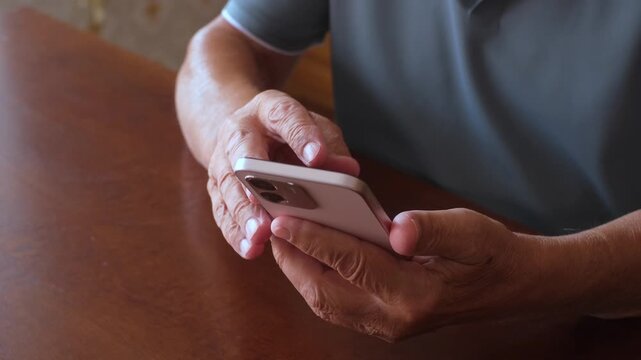 Closeup view of an older man's wrinkled hands holding and using a modern smartphone, scrolling and tapping the screen while sitting at a wooden table, representing technology for seniors