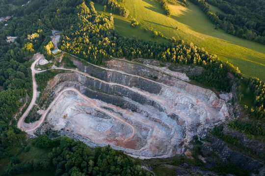 Aerial view of a large open pit stone quarry mine with terraced rock layers surrounded by green landscape during the sunset