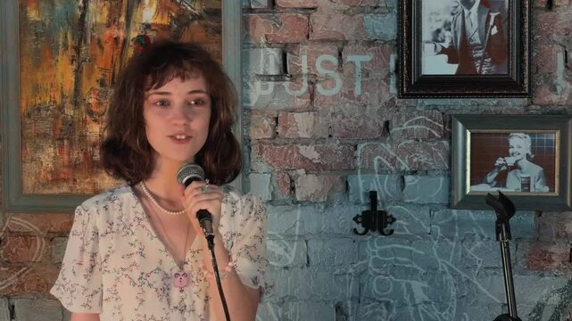 Cheerful young woman with curly brown hair in floral dress holding microphone telling funny story on stage at cozy retro club with painted brick wall background and vintage framed art portrait.