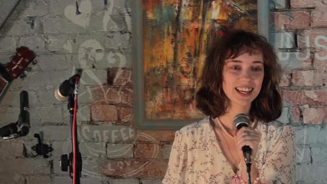 Charismatic young woman in a floral dress hosts an open mic event in a cozy indie cafe standing by a brick wall with vintage art holding a smartphone and microphone engaging the live audience