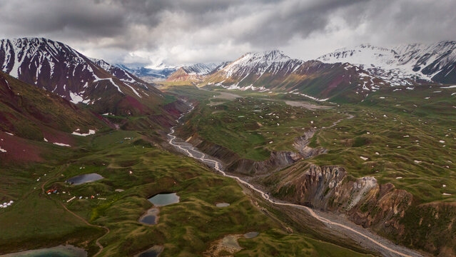 Aerial view of Tulpar Lake surrounded by rolling green hills, a winding river, and snow-capped mountains under a cloudy sky in Alay District, Osh Region, Kyrgyzstan.