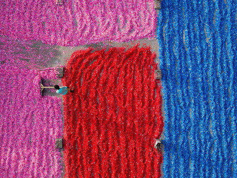 Aerial view of a worker raking vibrant pink, red, and blue plastic scraps spread across a large concrete surface in Dhaka, Dhaka Division, Bangladesh.