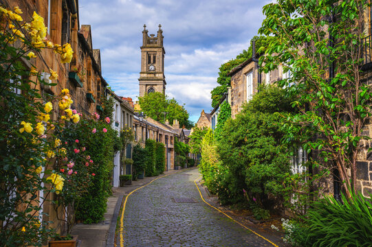 Cobblestone Circus Lane in Stockbridge, Edinburgh, lined with stone cottages, roses, and greenery, leading toward St Stephen's Church tower in Scotland, United Kingdom.