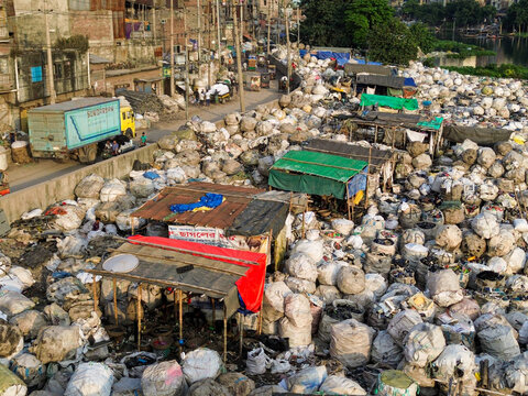 Aerial view of the Buriganga river canal area in Dhaka, showing numerous large white plastic waste bags piled near makeshift stalls and a road with a truck in Dhaka, Dhaka Division, Bangladesh.