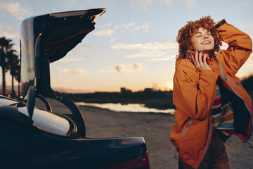 Naklejka premium woman, curly, hair, jacket, car, trunk, smile, sunset, lifestyle, outdoor, young woman closes car trunk while chatting and laughing outdoors, warm golden sky, travel mood today
