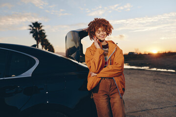 Naklejka premium Woman lifestyle outdoor portrait at sunset near car at beach lot, curly hair, orange jacket, relaxed smile, leaning beside vehicle, travel vibe in warm golden light with evening sky
