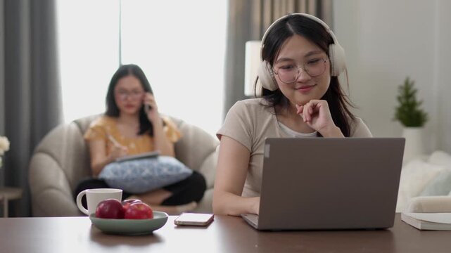 A young woman focuses on a laptop while working from home, with another woman relaxing in the background. Suitable for remote work, productivity, freelancing, digital business, and modern lifestyle