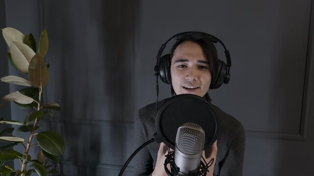 Expressive young Asian male artist wearing headphones passionately reciting classic poetry into condenser microphone with dramatic hand gestures in dark modern home recording studio medium shot