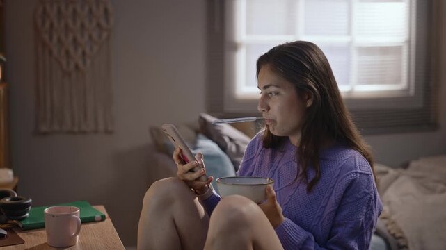 Young adult Caucasian woman eating from bowl while staring blankly at smartphone with spoon in mouth. Clip illustrates digital burnout and screen addiction for mental health campaigns