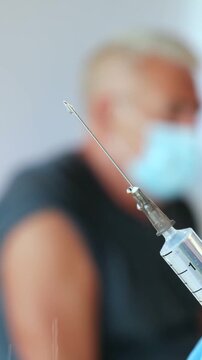 Macro view of medical liquid drop on steel needle tip of plastic syringe in vertical orientation, nurse hand preparing dose before blurred old patient vaccination. Clinical drug dosage preparation