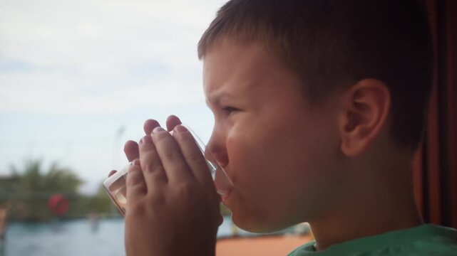 Child drinking water from a glass by a swimming pool, enjoying a calm outdoor moment with family during a summer day.