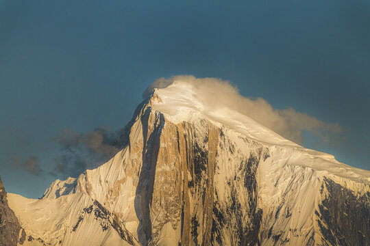 View of the snow-capped Spantik peak illuminated by golden hour light with dramatic rocky textures and wispy clouds in Nagar, Pakistan.
