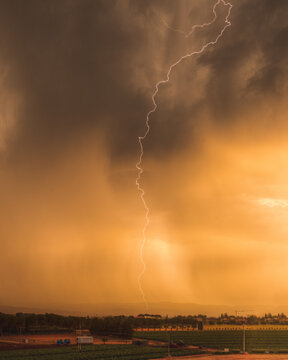 View of a dramatic lightning strike illuminating dark storm clouds over rural fields at sunset, Corridonia, Marche, Italy.