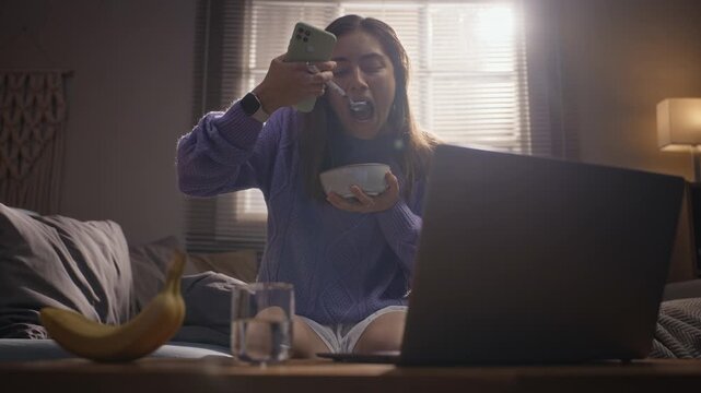 Young adult Caucasian woman eating breakfast while scrolling on smartphone with laptop open in foreground. Ideal for illustrating digital burnout, screen addiction or multitasking