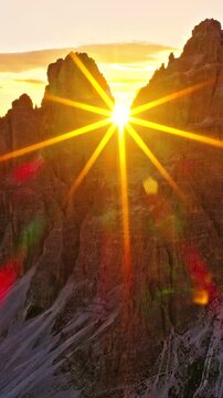 Aerial drone flight toward the glowing sunset between the iconic Tre Cime peaks in the Italian Dolomites. Dramatic alpine landscape with golden sunstar, lens flare and towering mountain cliffs.