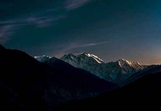 View of the snow-capped peak of Nanga Parbat rising above dark mountain silhouettes under a twilight sky in Pakistan.