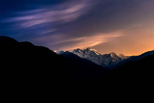 View of Nanga Parbat peak illuminated by the golden light of sunset against a dramatic purple and orange sky with silhouetted mountain ridges in the foreground in Pakistan.