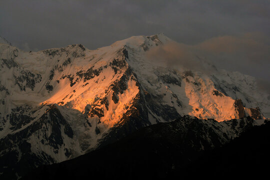 View of Nanga Parbat with snow-covered slopes illuminated by the warm golden light of sunset against a dark sky in Pakistan.