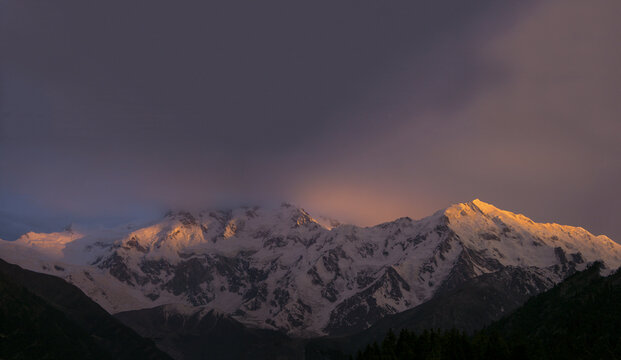 View of Nanga Parbat mountain peaks illuminated by golden hour light with snow-covered slopes and dark storm clouds in the sky in Chilas, Pakistan.