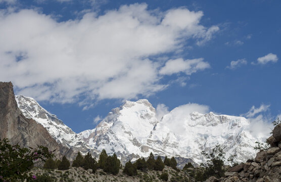 View of the snow-capped Nanga Parbat mountain peak rising above rocky terrain and green shrubs under a bright blue sky with scattered clouds in Chilas, Pakistan.
