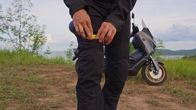 Rider Removes Yellow Knee Armor Pad From Protective Motorcycle Pants, Beside Parked Scooter Outdoors, Demonstrating Impact Protection and Rider Preparation