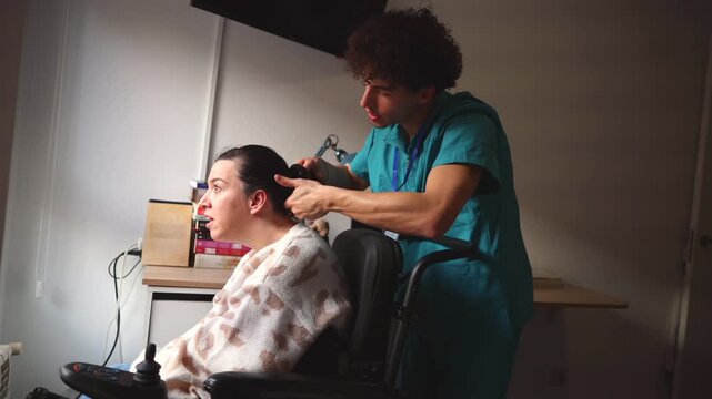 Caring male nurse helping a woman with a disability in a wheelchair, gently tying her hair into a ponytail as part of her daily personal care routine in a softly lit room at home or in a clinic