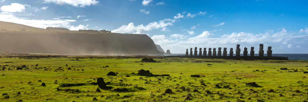 View of the iconic Moai statues at Ahu Tongariki standing in a row against the ocean with a grassy field and coastal cliffs under a bright blue sky in Easter Island, Valparaiso, Chile.