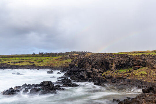 View of the rugged volcanic coastline with crashing waves, green grassy fields, and a faint rainbow in the sky at Easter Island, Valparaiso, Chile.