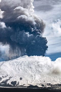 View of Mount Etna erupting with a massive volcanic ash plume rising above snow-covered slopes in Bronte, Sicily, Italy.