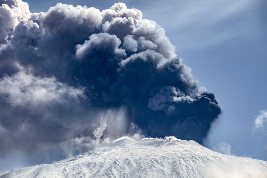 View of Mount Etna erupting with a massive plume of volcanic ash and smoke rising from the snow-covered summit into the blue sky in Bronte, Sicily, Italy.