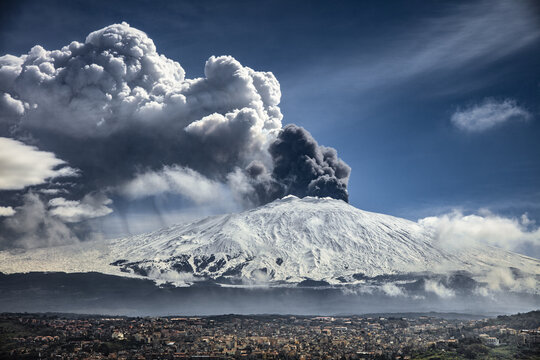 View of Mount Etna erupting with a massive volcanic ash plume rising above snow-covered slopes, overlooking the town of Bronte, Sicily, Italy.