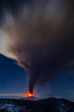 View of Mount Etna erupting at night with a glowing lava plume rising into the dark sky above the volcanic landscape in Catania, Sicily, Italy.