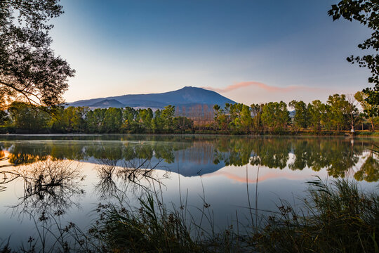View of Mount Etna reflecting in the calm waters of a lake at sunset, with lush trees and reeds along the shore in Catania, Sicily, Italy.