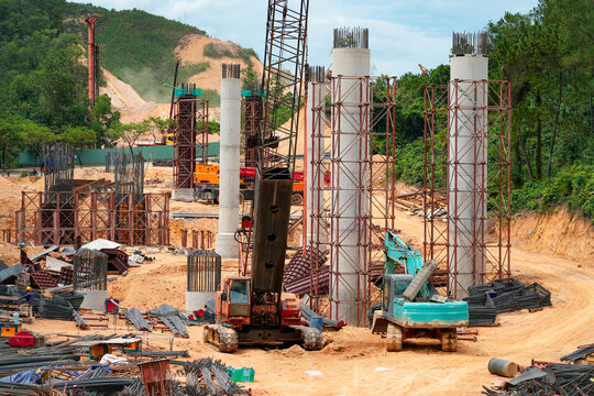Heavy construction work continues as workers prepare concrete columns for a new highway bridge project surrounded by lush green hills during the day.